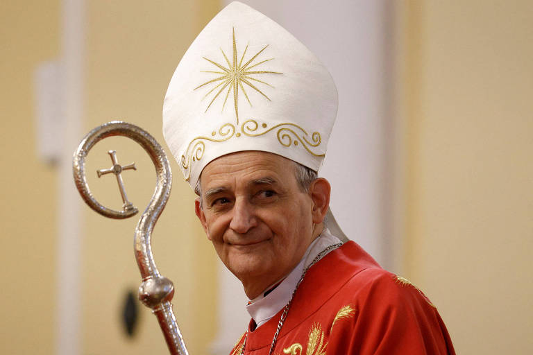 Um cardeal está vestido com uma túnica vermelha e um grande chapéu branco, adornado com um desenho de estrela. Ele segura um cajado pastoral prateado, que tem uma cruz na parte superior. O fundo é de um ambiente interno, com paredes em tons claros.