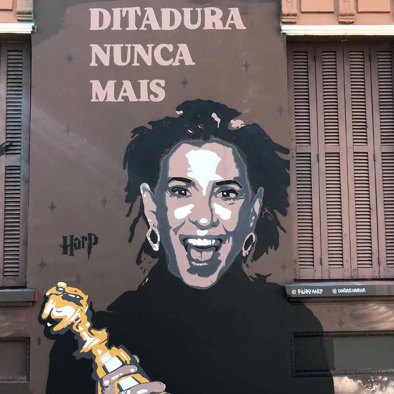 A imagem mostra um mural em uma parede， retratando uma mulher com cabelo cacheado e brincos grandes， sorrindo. Ela segura um troféu dourado. Acima da imagem， está escrito 039;DITADURA NUNCA MAIS039; em letras grandes e em destaque. O artista é identificado como 039;Harp039;.