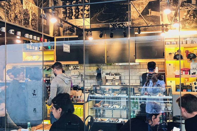 A imagem mostra o interior de uma cafeteria com várias pessoas sentadas em mesas. Há um balcão de atendimento ao fundo, onde um funcionário está preparando bebidas. As paredes são decoradas com fotos e há prateleiras com produtos expostos. A atmosfera parece acolhedora e moderna.