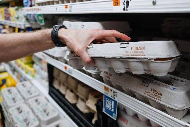 A imagem mostra uma mão segurando uma embalagem de ovos em uma prateleira de supermercado. A embalagem é de papelão e contém uma dúzia de ovos. Ao fundo， há outras embalagens de ovos e produtos alimentícios nas prateleiras.