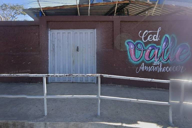 A imagem mostra a fachada de um edifício com uma porta de metal branca. Ao lado da porta， há um grafite colorido que diz 039;CED Valle do Amanhecer039;. A parede é de cor marrom e há uma grade de proteção na frente， com uma calçada de concreto. O céu está claro ao fundo.