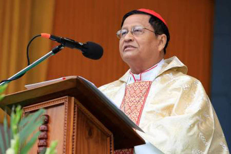 Um cardeal está em pé atrás de um púlpito, fazendo um discurso. Ele usa uma vestimenta religiosa dourada com detalhes vermelhos e óculos. O fundo é de cor laranja e há um microfone na frente dele. Plantas verdes estão visíveis na parte inferior da imagem.