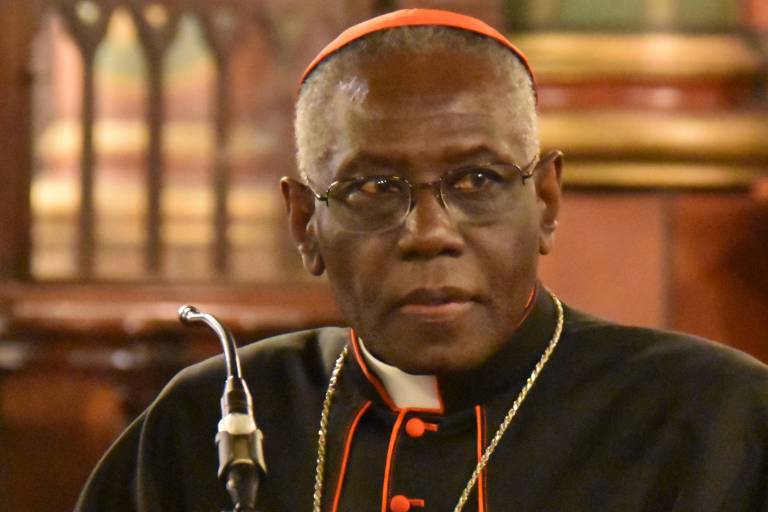 Um cardeal está sentado em uma mesa, olhando para a frente. Ele usa vestes religiosas pretas com detalhes em vermelho e um colarinho branco. Há um microfone à sua frente e um fundo de madeira com detalhes arquitetônicos visíveis.