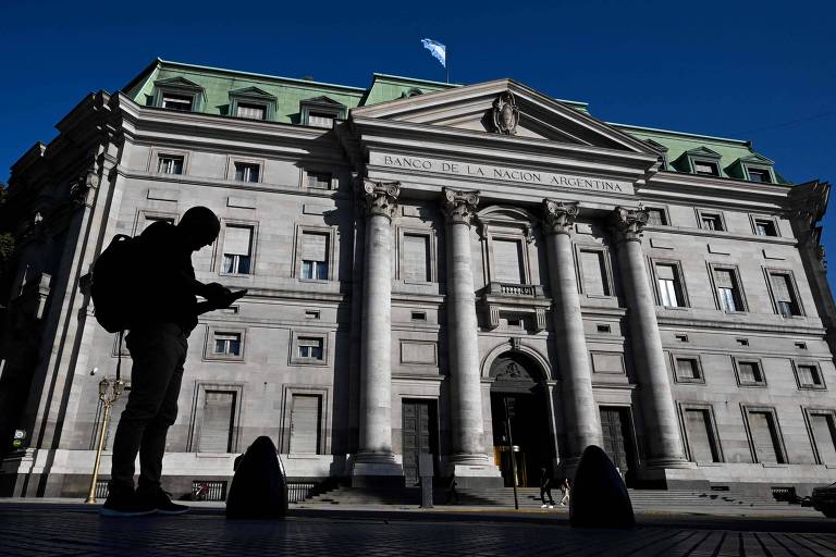 A imagem mostra a fachada do Banco de la Nación Argentina， um edifício imponente com colunas e detalhes arquitetônicos. À frente， uma silhueta de uma pessoa com uma mochila está olhando para um dispositivo móvel. O céu está claro e azul， e uma bandeira está visível no topo do edifício.