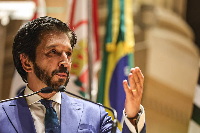 Um homem com cabelo escuro e barba está falando em um microfone. Ele usa um terno azul e uma gravata azul clara. Ao fundo， há bandeiras do Brasil e de outros países. O homem gesticula com a mão direita enquanto se expressa.