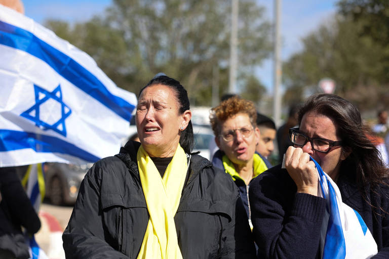 A imagem mostra um grupo de mulheres em um protesto， algumas delas chorando. Elas seguram bandeiras de Israel， que têm listras azuis e uma estrela de Davi. O ambiente parece ser ao ar livre， com árvores ao fundo e um céu parcialmente nublado.