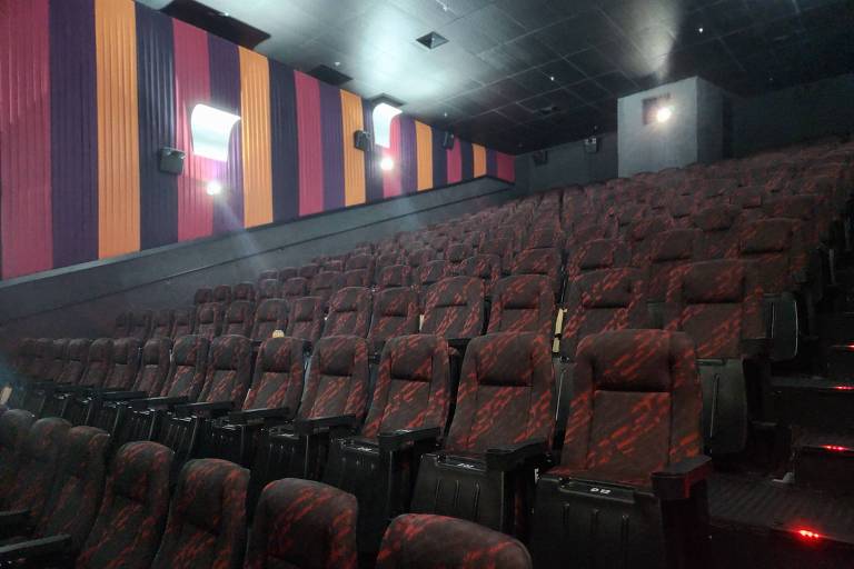 A imagem mostra o interior de uma sala de cinema com várias fileiras de poltronas vermelhas e pretas. As paredes são decoradas com painéis verticais em cores roxas, laranjas e amarelas. O ambiente é escuro, com algumas luzes de iluminação suave visíveis.