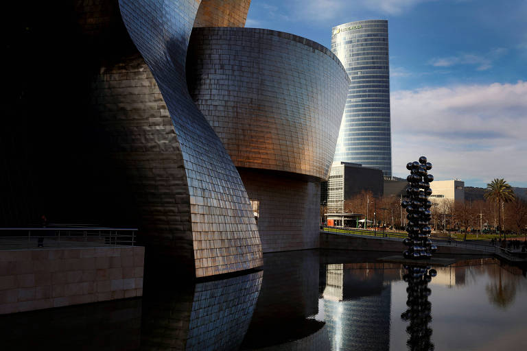 A imagem mostra a arquitetura do Museu Guggenheim de Bilbao， com suas formas curvas e superfícies metálicas refletindo a luz. Ao fundo， um edifício moderno de vidro se ergue， e à frente， há uma escultura em forma de coluna refletida na água calma de um lago próximo.