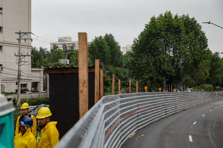 A imagem mostra uma área de construção em um ambiente urbano. Há uma cerca de proteção em primeiro plano， com trabalhadores usando capacetes amarelos. Ao fundo， é possível ver árvores e edifícios. O céu está nublado， indicando um clima possivelmente chuvoso.