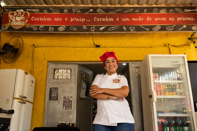 Uma mulher sorridente está em pé em frente a um restaurante， vestindo um uniforme de cozinheira e um lenço vermelho na cabeça. Ao fundo， há uma parede amarela com uma faixa que diz: 039;Cozinhar não é serviço... Cozinhar é um modo de amar as pessoas.039; Também é visível uma geladeira com bebidas e um relógio na parede.