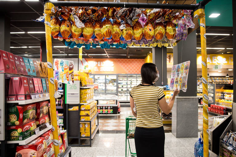 A imagem mostra uma pessoa de costas em um supermercado， em frente a um arco decorado com doces. O ambiente é bem iluminado， com prateleiras cheias de produtos variados， incluindo alimentos e bebidas. Ao fundo， há uma seção de congelados visível.