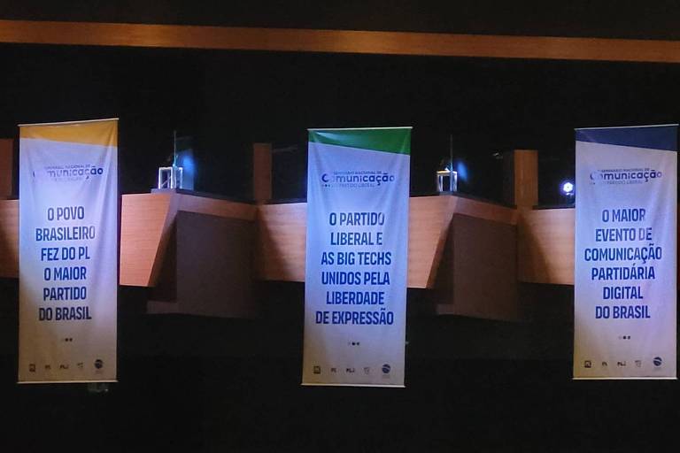 A imagem mostra três banners pendurados em um evento. Os banners têm fundo branco e texto em azul e preto. O texto é parcialmente legível， mas menciona temas relacionados a 039;comunicação039; e 039;educação039;. O ambiente parece ser um auditório ou sala de conferências， com mesas e iluminação suave.