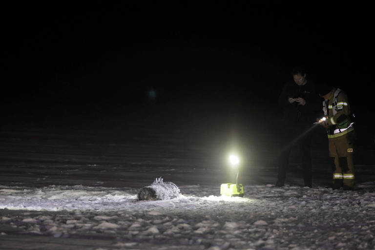 A imagem mostra uma cena noturna em um ambiente coberto de neve. À esquerda， há um objeto coberto de gelo no chão. À direita， duas pessoas estão presentes， uma delas segurando um celular e a outra usando um equipamento de proteção， iluminando a área com uma lanterna. O ambiente é escuro， com pouca visibilidade， exceto pela luz da lanterna.