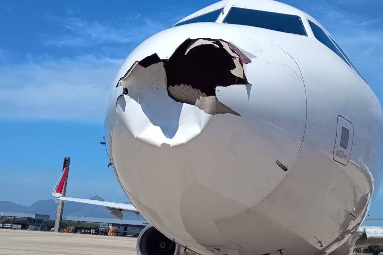 A imagem mostra a parte frontal de um avião com um grande buraco na fuselagem. O céu está claro e azul ao fundo， e a pista do aeroporto é visível. Há um trabalhador próximo ao avião， e outro avião pode ser visto ao fundo.