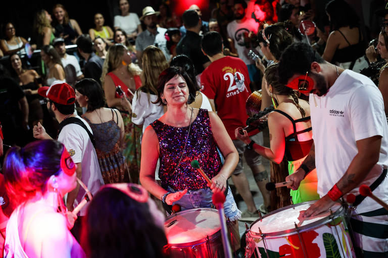 A imagem mostra um grupo de pessoas em uma festa animada， com foco em uma mulher tocando um tambor. Ela estaacute; sorrindo e vestindo uma blusa brilhante. Ao fundo， haacute; uma multidatilde;o de pessoas danccedil;ando e se divertindo， com algumas usando fantasias e acessoacute;rios coloridos. A iluminaccedil;atilde;o eacute; suave， com tons de vermelho， criando um ambiente festivo.