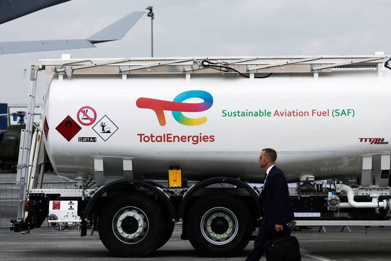 A imagem mostra um tanque de combustível de aviação sustentável da TotalEnergies， com o logotipo da empresa e a inscrição 039;Sustainable Aviation Fuel (SAF)039;. O tanque está montado em um caminhão e um homem de terno caminha ao lado dele. Ao fundo， é possível ver uma aeronave e um céu nublado.