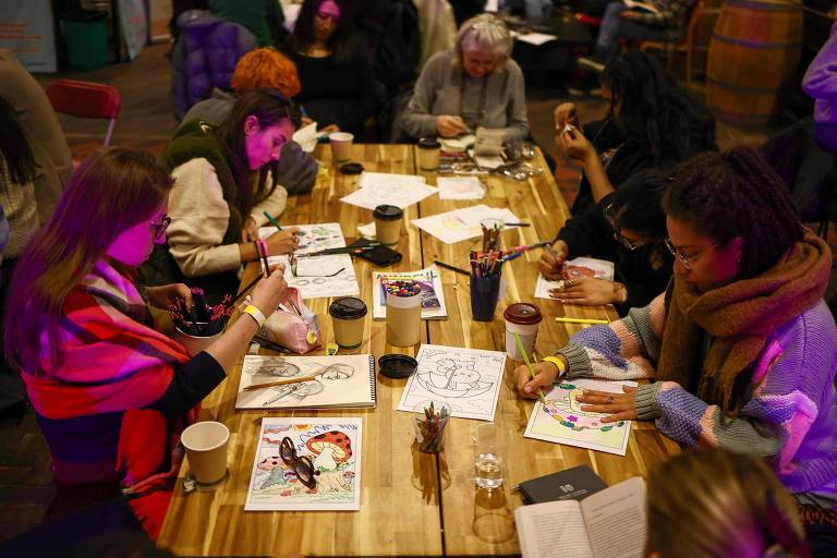 A imagem mostra um grupo de pessoas sentadas ao redor de uma mesa de madeira， envolvidas em uma atividade de colorir. Elas estão usando canetas coloridas e lápis para colorir desenhos em folhas de papel. Algumas pessoas estão segurando copos de bebida， enquanto outras têm materiais de arte ao seu redor. O ambiente é iluminado com luzes suaves e há barris de madeira ao fundo.