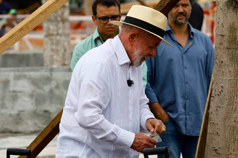 A imagem mostra um homem de cabelo grisalho， usando uma camisa branca e um chapéu claro， que está se inclinando para baixo， aparentemente manipulando algo em suas mãos. Ao fundo， há dois homens observando， um deles com óculos e uma camisa azul. O ambiente parece ser um local de evento ao ar livre， com estruturas de madeira e uma área de construção visível.
