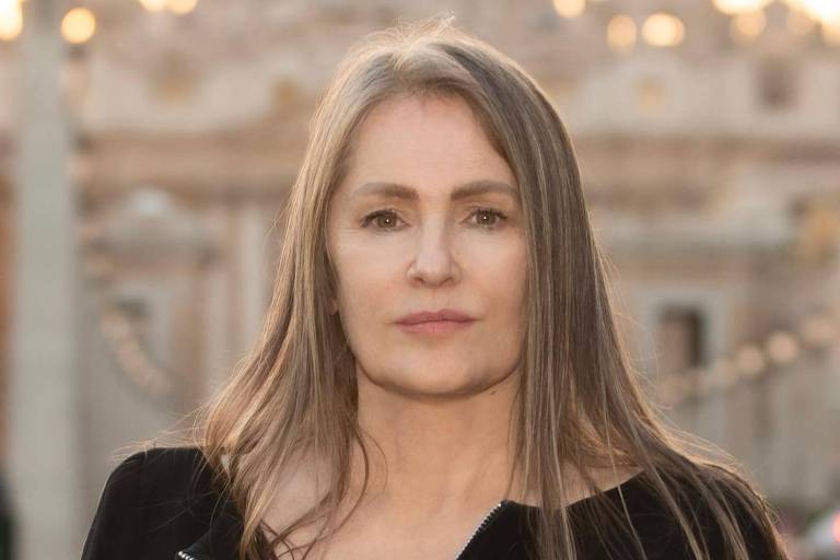 Uma mulher com cabelo longo e liso, usando uma blusa preta com zíper, está em pé em um local ao ar livre. Ao fundo, pode-se ver uma grande estrutura arquitetônica, possivelmente uma catedral ou igreja, com cúpulas e detalhes ornamentais. A luz do sol está suave, indicando um momento do dia próximo ao pôr do sol.