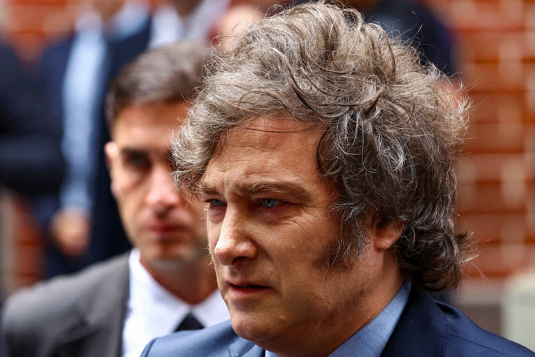 A imagem mostra um homem com cabelo cacheado e desgrenhado， usando um terno escuro e uma camisa clara. Ele parece estar em um ambiente externo， com um fundo desfocado que sugere a presença de outras pessoas. O homem tem uma expressão séria e está olhando para baixo， enquanto um outro homem， com cabelo curto e terno claro， aparece ao fundo.