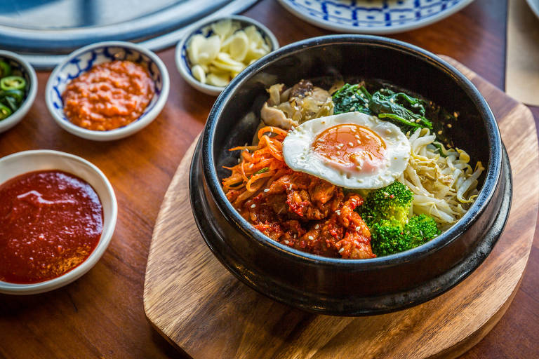 A imagem mostra um prato de bibimbap servido em uma tigela de pedra quente. O prato contém arroz, vegetais variados, carne e um ovo frito por cima. Ao fundo, há pequenos pratos com acompanhamentos, incluindo molhos e vegetais. A mesa é de madeira e há um ambiente iluminado ao fundo.