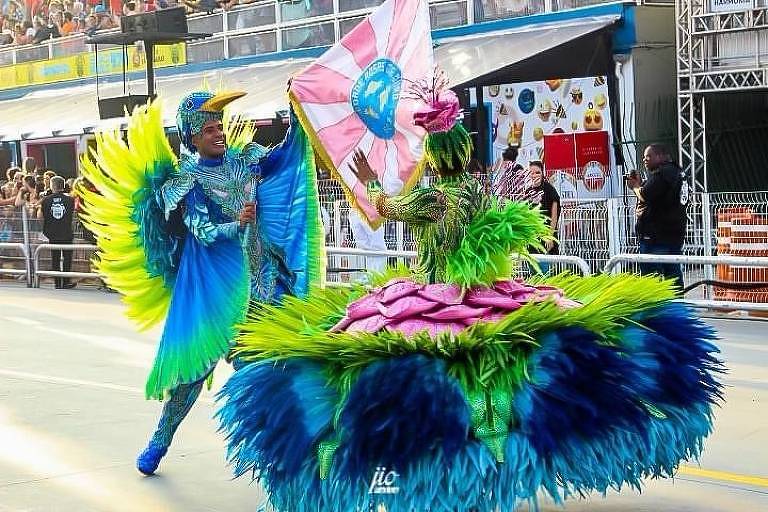 A imagem mostra duas pessoas dançando em um desfile de Carnaval. Uma delas está vestida com um traje colorido que imita penas de pássaro, predominantemente azul e verde. A outra pessoa usa um vestido volumoso, também em tons de verde, com detalhes em rosa. Ambas seguram uma bandeira colorida enquanto se apresentam em uma passarela, com uma multidão ao fundo.