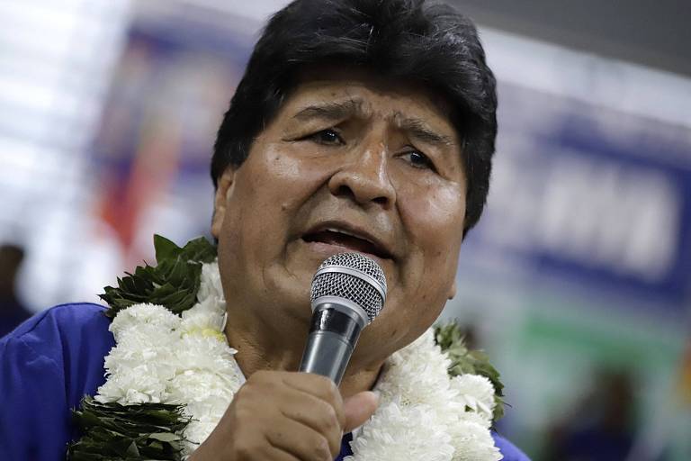 Um homem de cabelo escuro e liso， usando uma camisa azul e um colar de flores brancas， está falando em um microfone. O fundo é desfocado， mas parece haver uma multidão e banners ao redor.