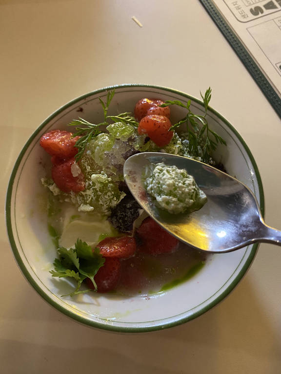 Uma tigela com uma mistura de ingredientes. Há morangos cortados, folhas verdes, um pouco de gelo e um molho verde em uma colher. O fundo da tigela é branco com uma borda verde.