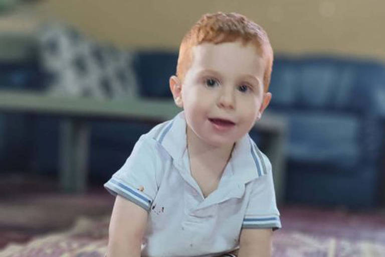 Um menino pequeno com cabelo ruivo e pele clara está sentado no chão， em um tapete. Ele usa uma camisa branca com detalhes e calças curtas. O menino sorri enquanto brinca com um brinquedo azul. Ao fundo， há um sofá azul e uma mesa.