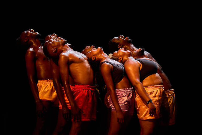 A imagem mostra um grupo de seis dançarinos em uma pose sincronizada, com os corpos inclinados para trás e as cabeças levantadas. Eles estão em um fundo escuro, com iluminação que destaca seus rostos e corpos. Os dançarinos usam shorts coloridos, variando entre tons de laranja e rosa, e estão em uma expressão de alegria ou liberdade.