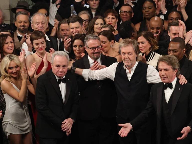 A imagem mostra um grande grupo de pessoas em um evento， a maioria aplaudindo. No centro， há quatro homens， um deles com um colete preto e uma camisa branca， e os outros em trajes formais. Ao fundo， várias pessoas estão sorrindo e aplaudindo， incluindo mulheres em vestidos elegantes. O ambiente parece ser um auditório ou um salão de eventos.