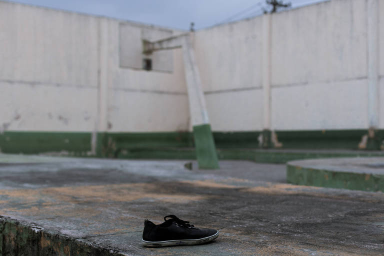 A imagem mostra um tênis preto solitário em uma superfície de concreto. Ao fundo， há uma parede branca com algumas aberturas e uma estrutura verde. O ambiente parece ser um espaço externo， possivelmente um campo ou área de recreação， com um céu nublado.