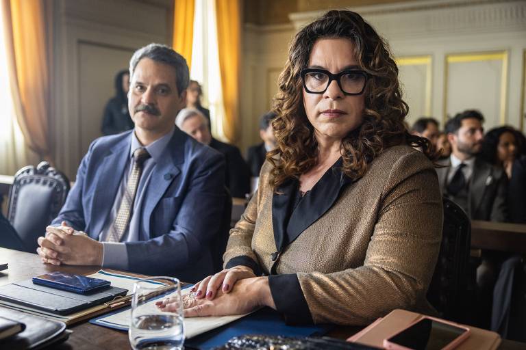 A imagem mostra um ambiente de tribunal com duas pessoas em primeiro plano. À esquerda, um homem com bigode e cabelo grisalho, vestido com um terno azul e gravata. À direita, uma mulher com cabelo cacheado e óculos, usando um blazer dourado. Ambos estão sentados à mesa, com documentos e um copo de água à frente. Ao fundo, outras pessoas estão sentadas, observando. A luz entra pelas janelas com cortinas amarelas.