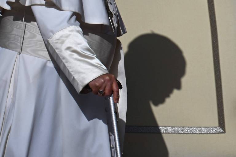 A imagem mostra uma mão segurando uma bengala， com uma figura vestida de branco ao fundo. A sombra da figura é projetada na parede， criando um contorno que sugere um rosto. A iluminação destaca a bengala e a vestimenta branca， enquanto a parede é de uma cor clara.