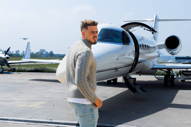 Um homem está caminhando em direção a um jato particular em uma pista de pouso. Ele usa um suéter claro e jeans, e está sorrindo. O céu está claro e ensolarado, com algumas nuvens. Ao fundo, há outros aviões estacionados.