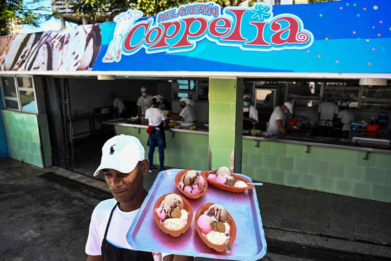 Um homem está segurando uma bandeja com sorvetes em uma loja chamada 039;Coppelia039;. O fundo mostra a fachada da sorveteria， que tem uma placa colorida com o nome 039;Coppelia039;. O homem usa uma camiseta branca e um boné. Na loja， várias pessoas estão trabalhando.