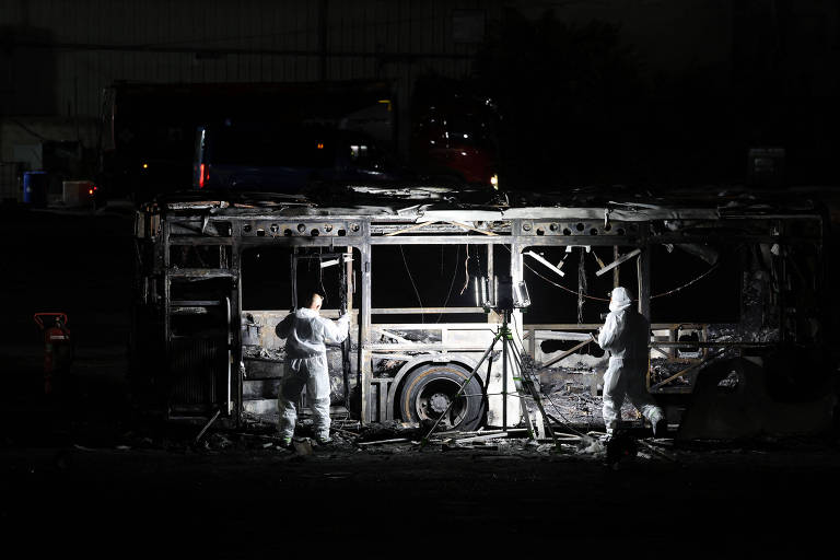 A imagem mostra um ônibus queimado em um ambiente escuro， com duas pessoas vestindo trajes de proteção branca， aparentemente realizando uma investigação. O ônibus está danificado， com a estrutura exposta e partes queimadas visíveis. Ao fundo， há um veículo não identificado e a iluminação é baixa， destacando a cena de destruição.