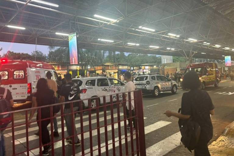 A imagem mostra uma cena urbana ao entardecer， com várias pessoas caminhando em uma faixa de pedestres. Há veículos de emergência， incluindo ambulâncias e carros de polícia， estacionados em uma área coberta. O ambiente é iluminado por luzes artificiais e há um painel publicitário visível ao fundo.