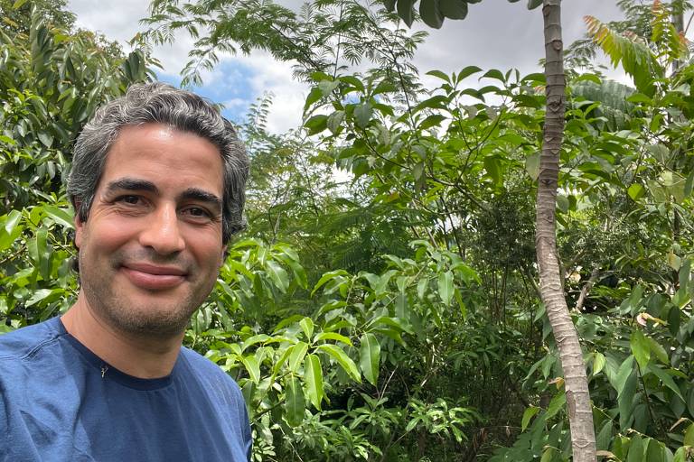 Um homem sorridente está posando para a foto em um ambiente natural， cercado por vegetação densa. Ele tem cabelo grisalho e usa uma camiseta azul. Ao fundo， há várias plantas verdes e um céu parcialmente nublado.