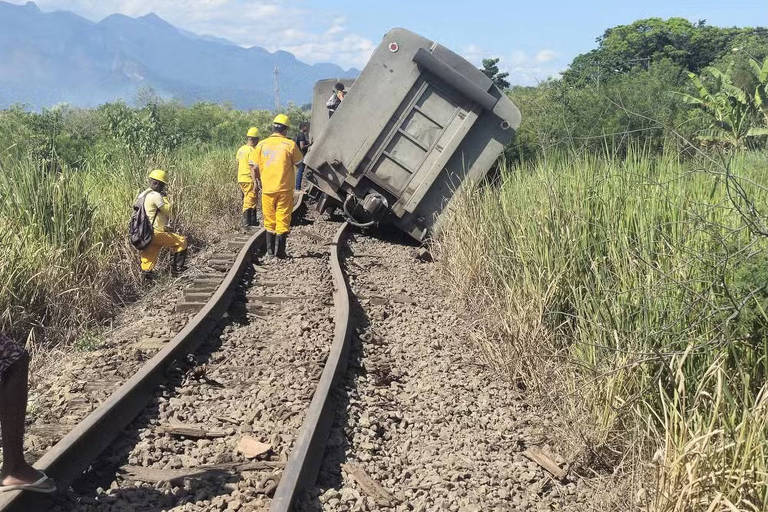 A imagem mostra um trem descarrilado em uma linha férrea. Um dos vagões está tombado para o lado， enquanto trabalhadores em uniformes amarelos estão próximos ao local do acidente. Ao fundo， há vegetação alta e montanhas sob um céu claro.