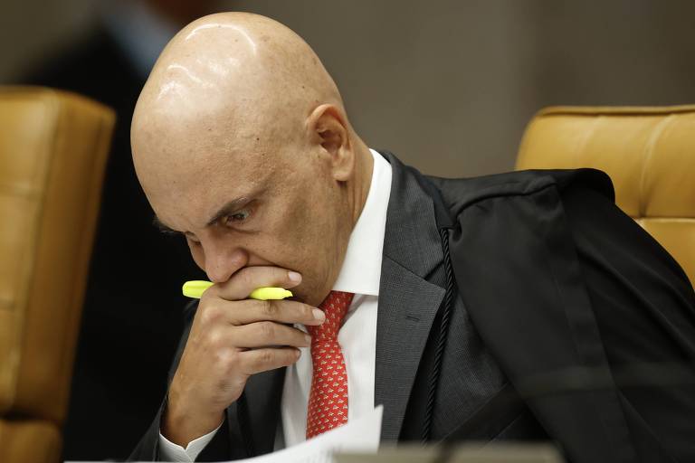 Um homem careca， vestido com um terno escuro e uma gravata vermelha， está sentado em uma cadeira. Ele está com a cabeça baixa， pensativo， segurando um marcador amarelo com a mão direita. O ambiente parece ser uma sala de tribunal， com cadeiras de couro ao fundo.