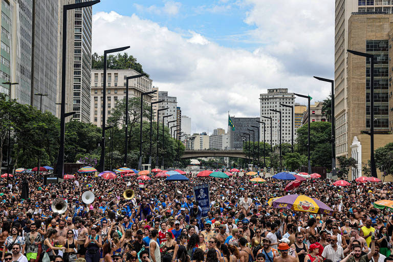 A imagem mostra uma grande multidão reunida em uma avenida urbana， cercada por prédios altos. O céu está parcialmente nublado， e há várias pessoas usando roupas coloridas. Algumas pessoas estão segurando guarda-sóis coloridos， e há um grupo de músicos ao fundo. A cena transmite um ambiente festivo e animado.