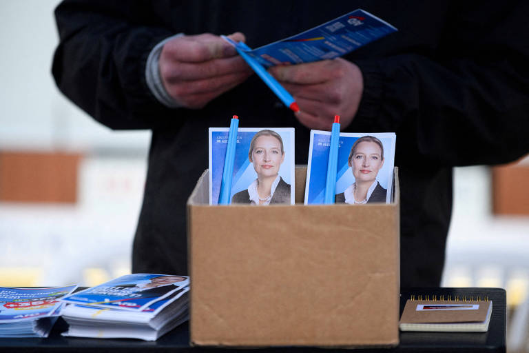 A imagem mostra uma pessoa segurando um folheto azul enquanto está em frente a uma caixa de papelão. Dentro da caixa， há várias canetas e fotos de uma mulher com um fundo azul. Também há folhetos espalhados sobre a mesa.