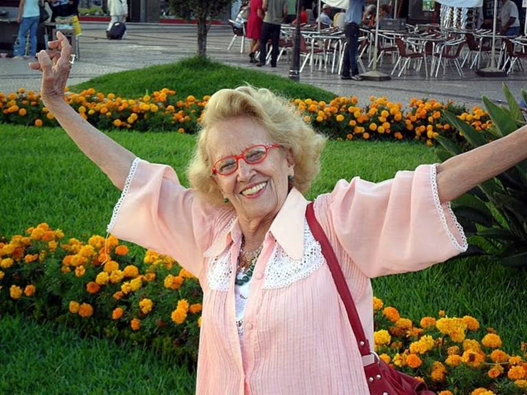 Uma mulher idosa com cabelo loiro e óculos， vestindo uma blusa rosa， está sorrindo e levantando os braços em um jardim. Ao fundo， há flores amarelas e um espaço com mesas e cadeiras.