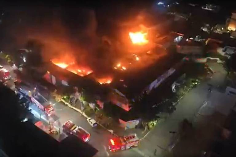 A imagem mostra um incêndio em uma fábrica de isolantes térmicos na Zona Sul de São Paulo. Chamas intensas e fumaça densa saem do telhado do edifício, enquanto viaturas de bombeiros estão estacionadas nas proximidades, prontas para combater o fogo. A cena é noturna, com iluminação artificial visível nas ruas ao redor.