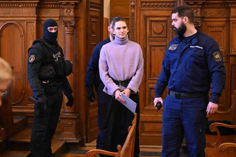 A imagem mostra uma cena em um tribunal. Uma pessoa com um suéter roxo está sendo acompanhado por dois agentes de segurança. Um dos agentes está vestido com um uniforme azul e segura um objeto, enquanto o outro está usando uma máscara preta e um colete à prova de balas. O ambiente é de madeira escura, com detalhes arquitetônicos visíveis ao fundo.