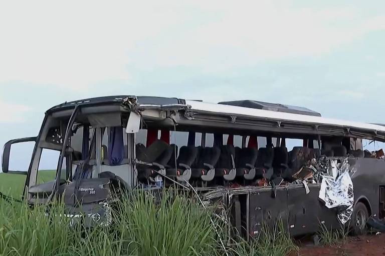 A imagem mostra um ônibus danificado， tombado em um campo. O veículo apresenta danos significativos na lateral， com janelas quebradas e parte da estrutura exposta. A vegetação ao redor é alta， e o céu está nublado， sugerindo um clima sombrio.