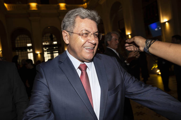 A imagem mostra um homem sorridente, vestido com um terno escuro e uma gravata vermelha, em um ambiente de evento social. Ele está em movimento, com os braços abertos, e parece estar interagindo com outras pessoas ao seu redor. O fundo é iluminado, com outras figuras indistintas ao longe.