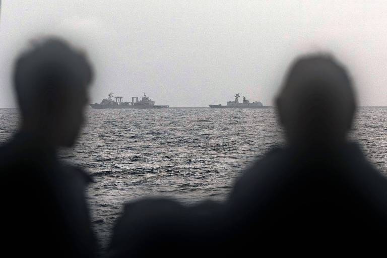 A imagem mostra a silhueta de duas pessoas em primeiro plano， com o mar ao fundo. No horizonte， são visíveis dois navios de guerra， com um céu nublado e uma atmosfera sombria.