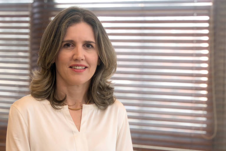 A imagem mostra uma mulher com cabelo longo e ondulado， usando uma blusa branca. Ela está sorrindo e posando para a câmera， com um fundo de janelas com persianas. A iluminação é suave e destaca seu rosto.